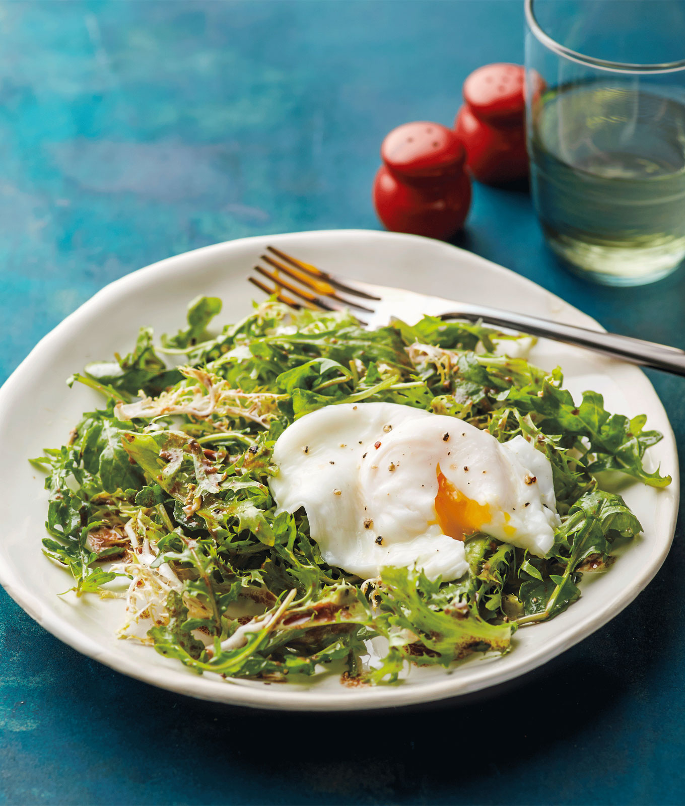 Low Sugar Breakfast Recipes - A poached egg on an arugula salad. It's on a white plate with a fork in it. It sits on a light teal surface. Next to the plate is a glass filled with a clear yellow liquid and red salt and pepper shakers.