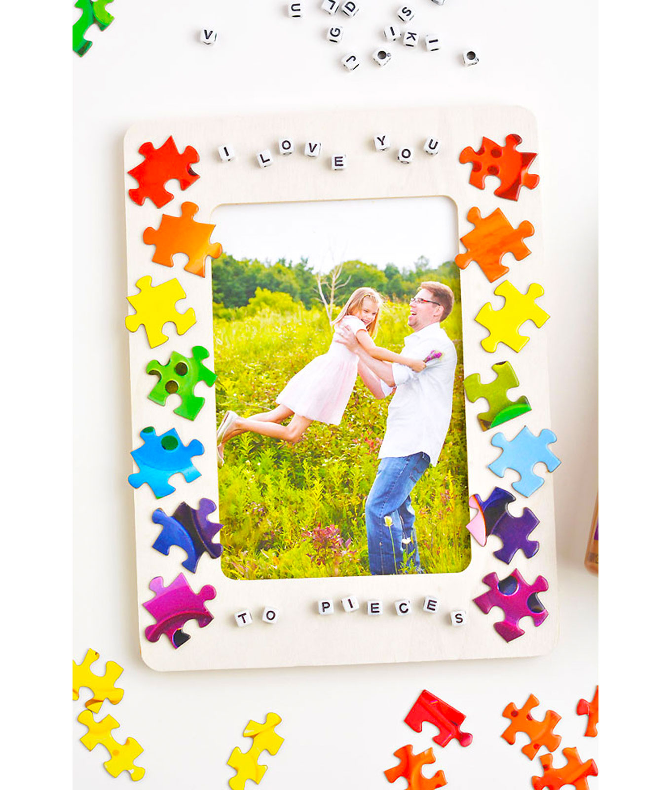 mothers-day-crafts: A white picture frame decorated with rainbow colored puzzle pieces in front of a white background. There are stray pieces next to the frame.