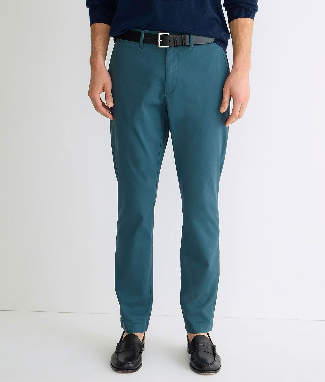 jcrew-mens-winter-collection: A man wears a pair of teal colored chino pants, a belt, black slide loafers and a dark shirt in front of a white background.
